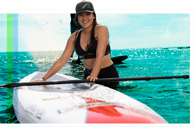A female student standing in the water next to a paddleboard with the oar laid across it.