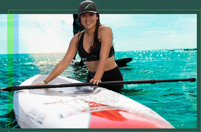 A female student standing in the water next to a paddleboard with the oar laid across it.