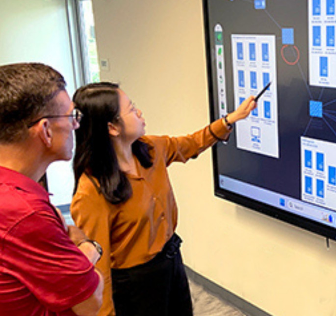 A professor standing next to a student pointing to a LED screen with infographics of computer systems.