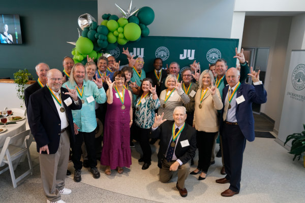 A group of people posing for photo in front of a JU panel ornated with green ballons.