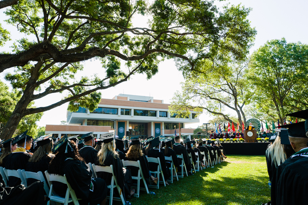 Photo taken behind a group of graduates sitting down during a morning graduation ceremony with JU's Howard Building in the background
