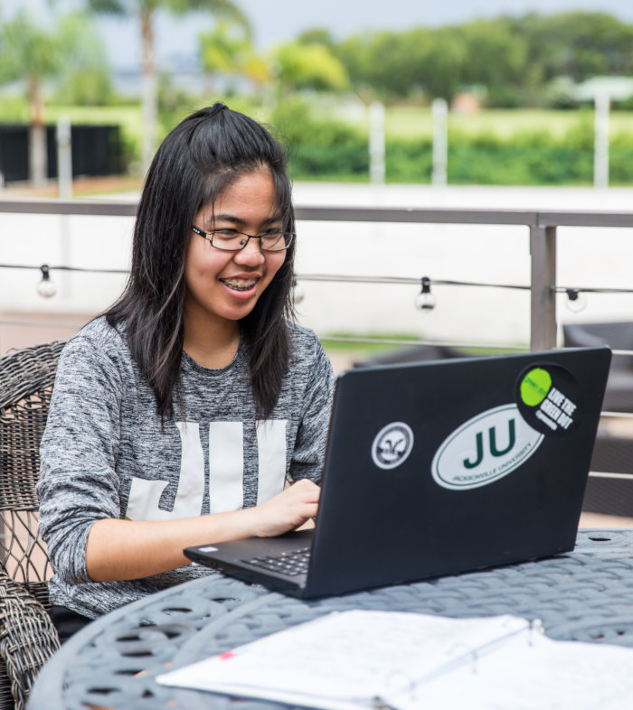 A student seating outdoors wearing a Jacksonville University t-shirt while working on their laptop. 