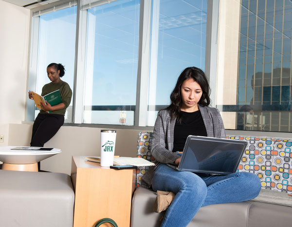 A young woman sits on a couch and works on a laptop. Another woman looks at her phone while leaning against the wall in the distant background.